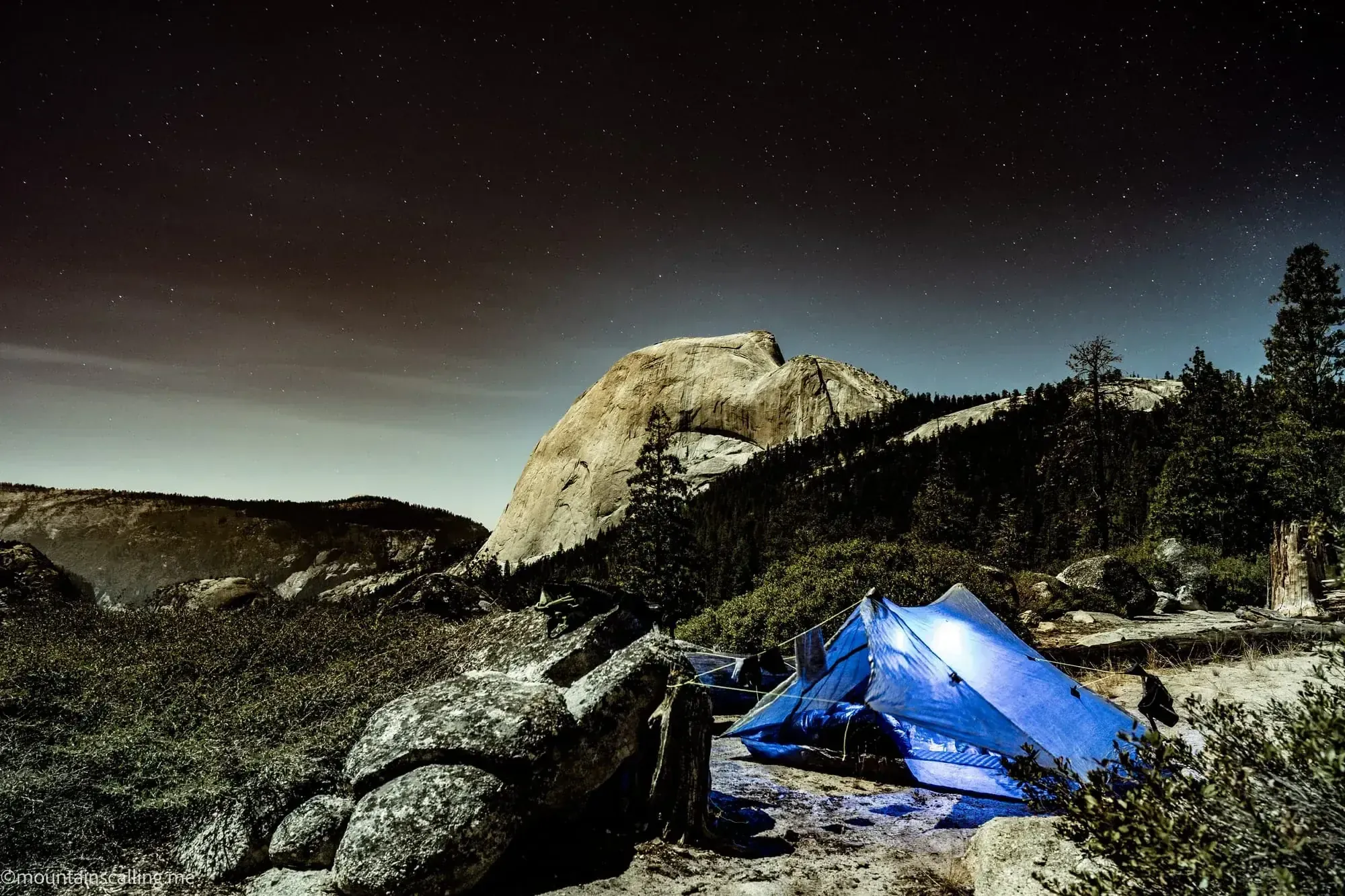 Blue tent under starry night sky with Half Dome granite formation illuminated in background at Yosemite backpacking camp