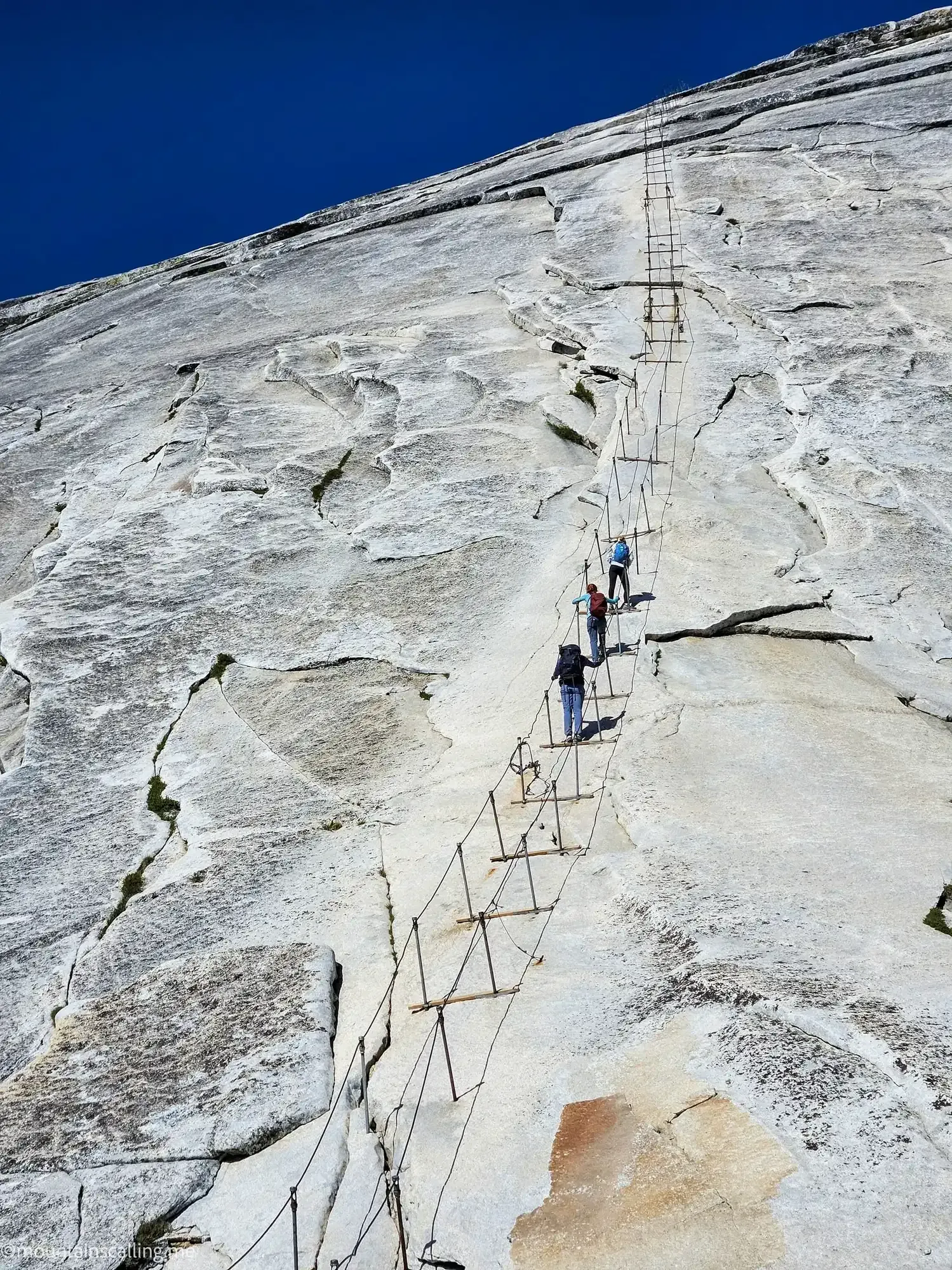 Hikers climbing Half Dome cables section with metal ladder system on steep granite face in Yosemite