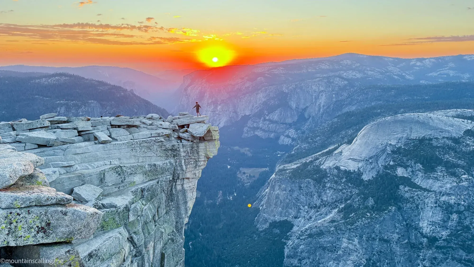 Hiker standing on Half Dome summit cliff edge at sunset overlooking Yosemite Valley granite peaks