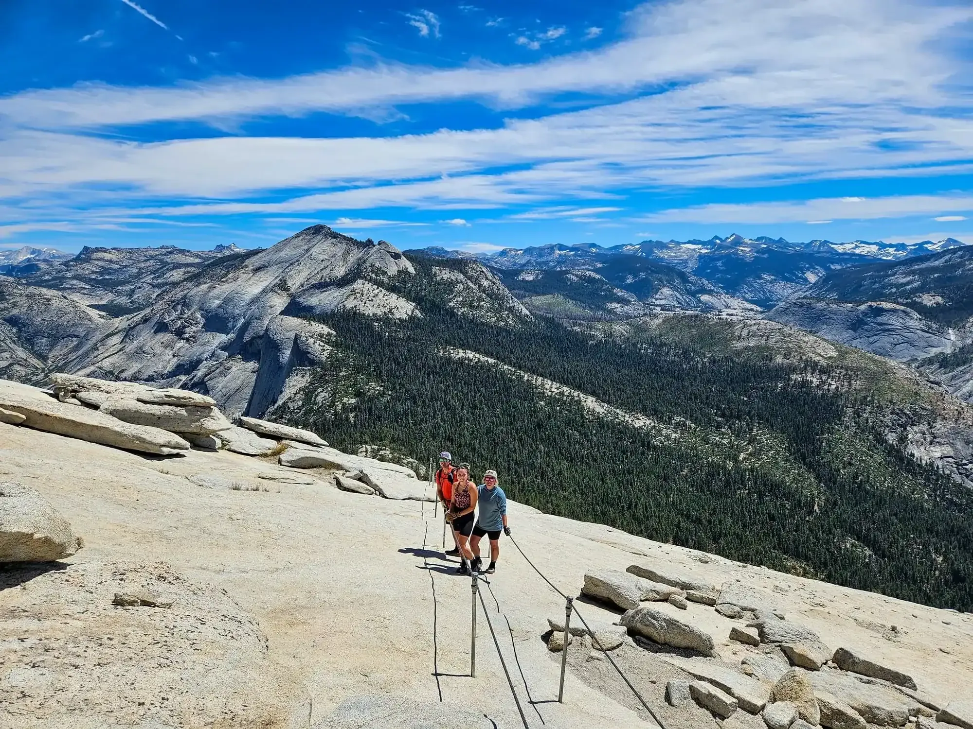 Three hikers celebrating on Half Dome summit with cables and Yosemite wilderness panorama behind them