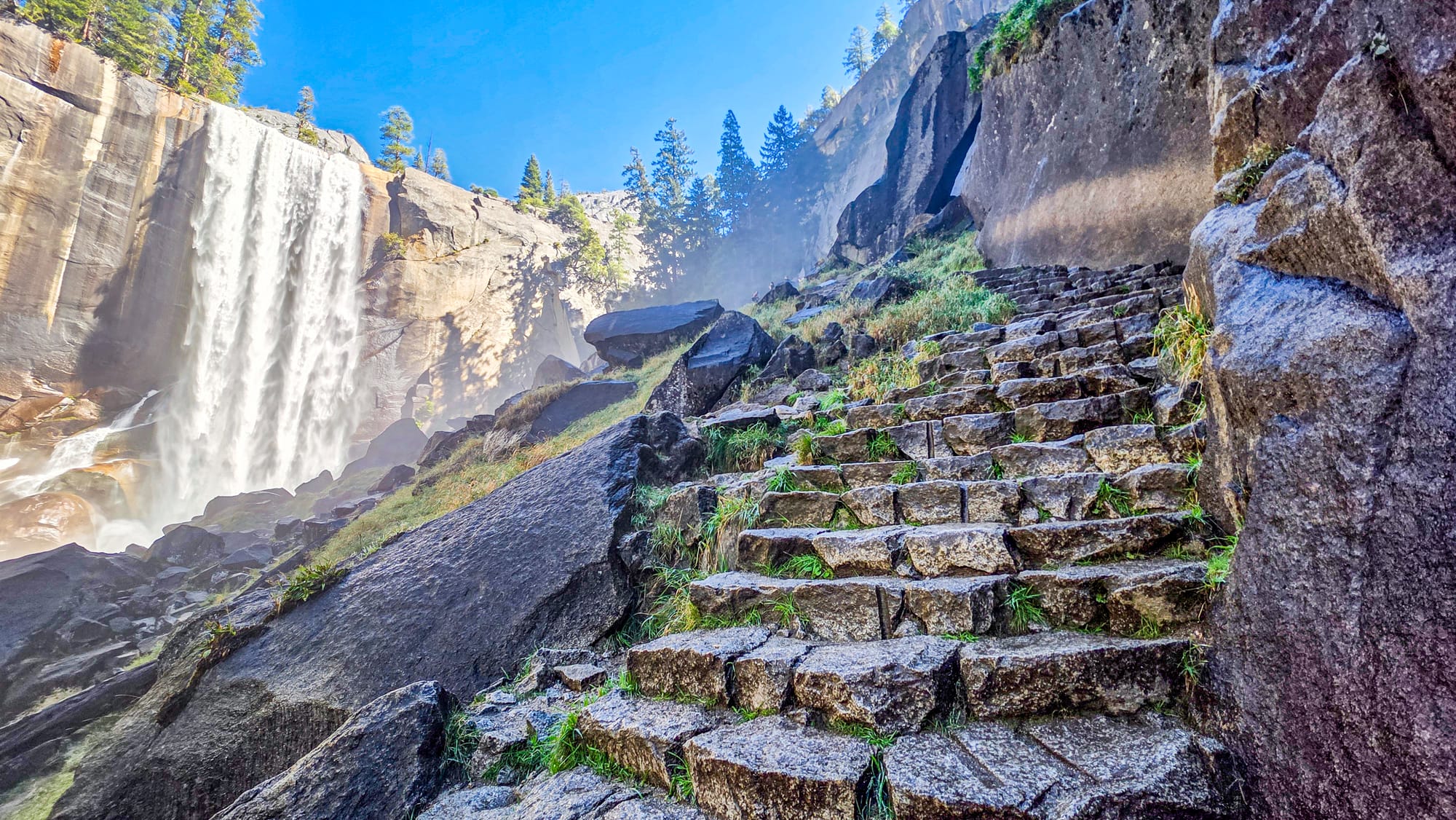Granite steps of the Mist Trail climbing alongside Vernal Fall in Yosemite National Park with mist and sunlight illuminating the waterfall | Yosemite Life | Yosemite.Life