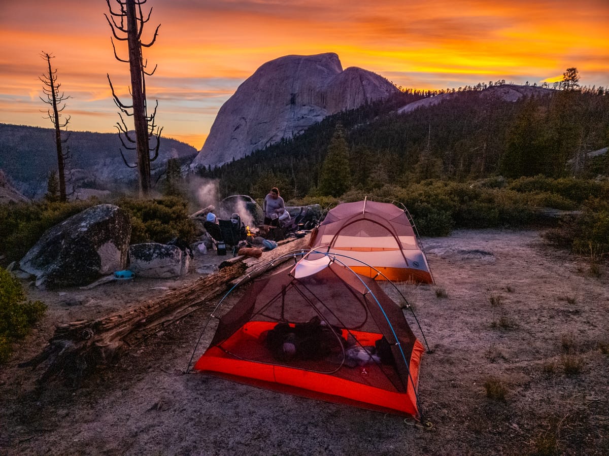 Backcountry campsite with two tents and campers gathered around a fire at sunset, Half Dome glowing orange in the background | Yosemite Life